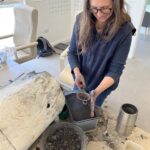 Jane wearing a blue sweater and apron, handling charcoal material with a metal scoop in a workspace.